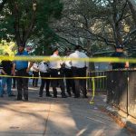 Police officers gather behind yellow tape on a tree-lined sidewalk