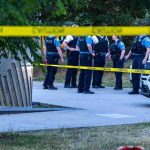 police officers stand behind caution tape at a park scene