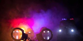 Gunfire: Agents Fired on During Drug Raid Handcuffs on a surface with police cars and flashing lights in smoky darkness