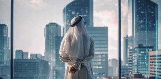 Man in traditional robe looking out office window at modern skyscrapers